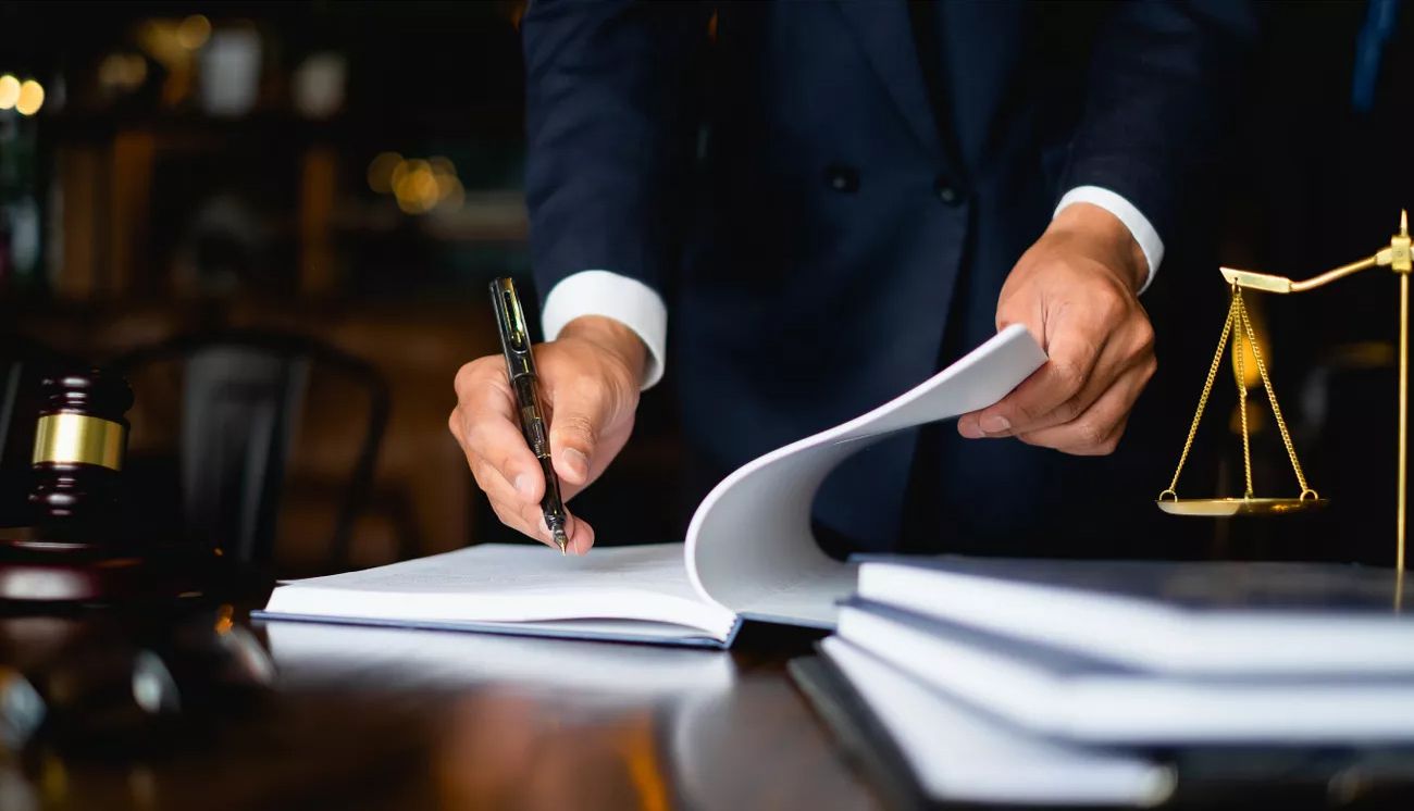 Eine Person unterzeichnet etwas in einem Buch, links daneben ist ein Holzhammer, rechts davon ist eine goldene Waage.